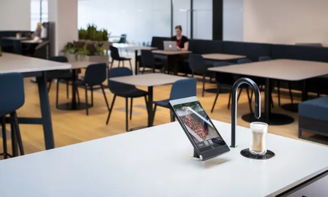 Matte black TopBrewer with deluxe iPad holder in the foreground and person working on laptop in background