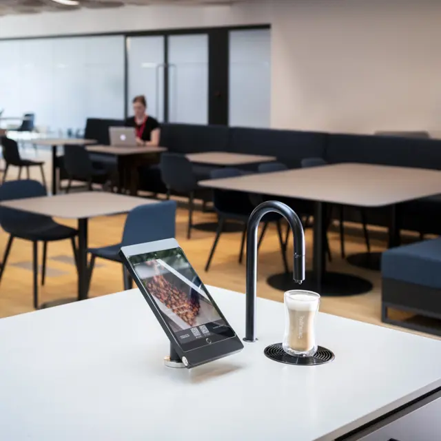Matte black TopBrewer with deluxe iPad holder in the foreground and person working on laptop in background