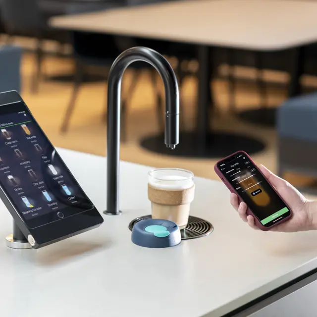 Image showing a matte black TopBrewer coffee machine with a reusable coffee cup under the swan neck and the TopBrewer app on an iPhone