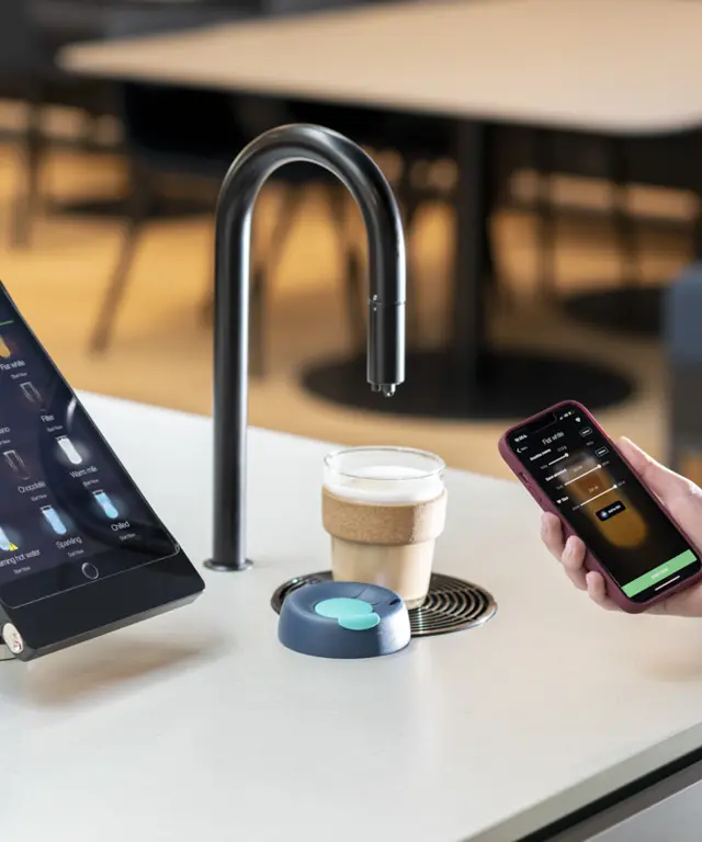 Image showing a matte black TopBrewer coffee machine with a reusable coffee cup under the swan neck and the TopBrewer app on an iPhone