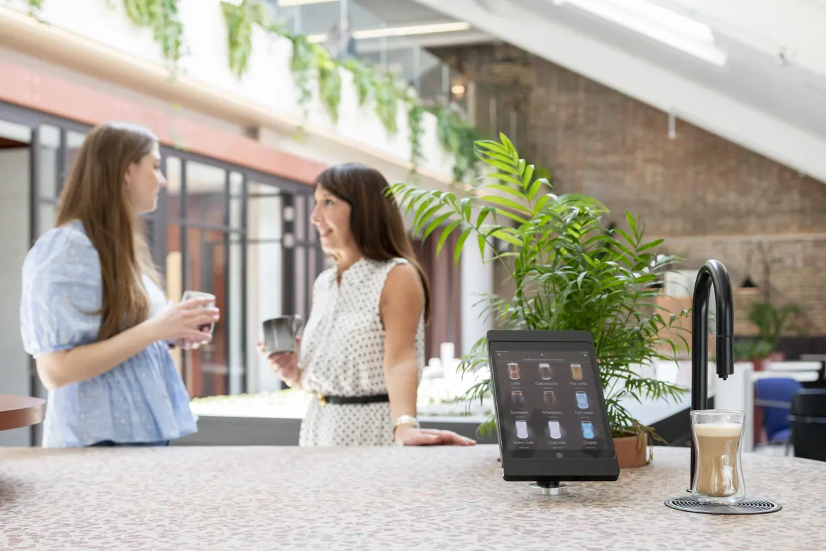 Image showing a matte black TopBrewer commercial coffee machine in the foreground and two people chatting in the background