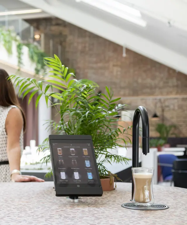 Image showing a matte black TopBrewer commercial coffee machine in the foreground and two people chatting in the background