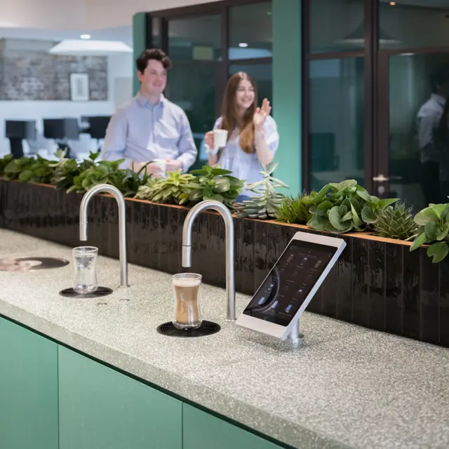 Image shows TopBrewer commercial coffee machine and TopWater built into a cabinet, with 2 people chatting in the background