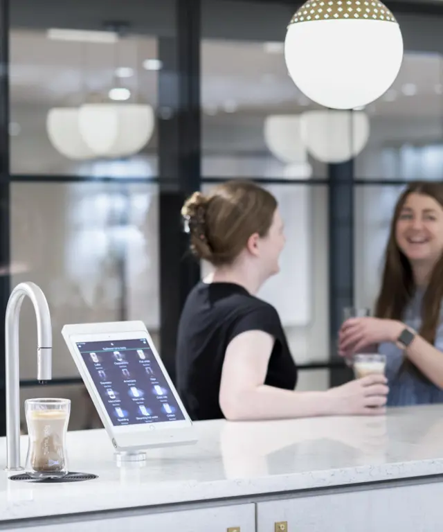 Image shows TopBrewer commercial coffee machine in the foreground with 2 colleagues chatting in the background