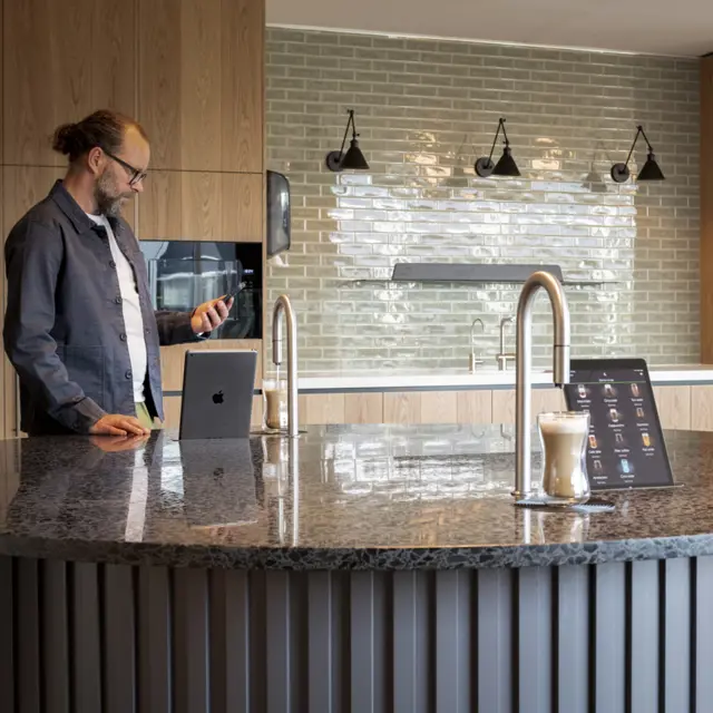2 TopBrewer coffee machines built into a circular island, with a man in the background looking at his phone